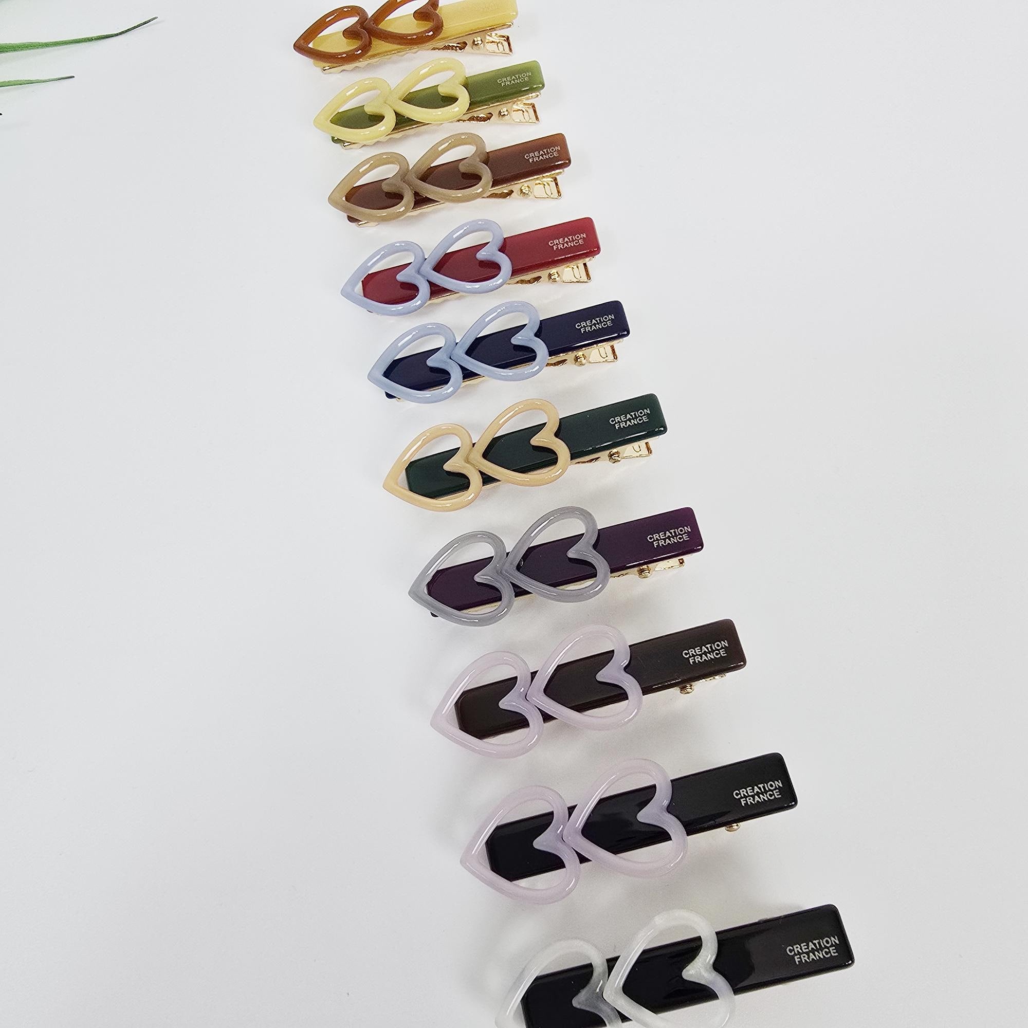 Cellulose acetate twin heart flat hair clips in pastel and tortoise shell colors for bangs and side hair. A collection of heart-shaped hair clips in various colors. The clips feature a double heart design and are attached to rectangular barrettes. Colors include yellow, brown, red, blue, green, purple, and white. Each clip has 'CREATION FRANCE' printed on it.